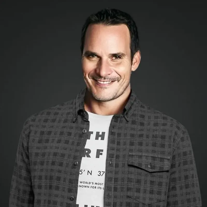 Portrait of Ellis Norman, a light-skinned man with dark hair, dark eyes, and a stubble beard, smiling against a dark gray background, dressed in a dark gray plaid button-up shirt over a white t-shirt with partially visible black text.