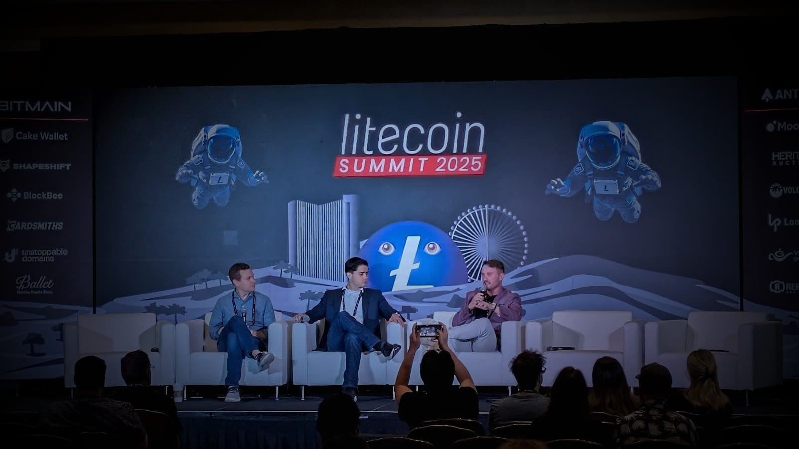 Three men seated in white chairs on stage during Litecoin Summit 2025, with a large screen behind them displaying the event title, floating astronaut mascots, and sponsor logos. One speaker holds a microphone and gestures as the audience records the panel.