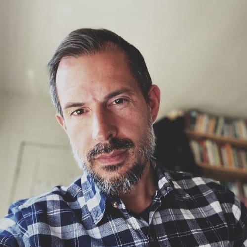 Philip Milestone - General Counsel at Valora, close-up portrait of a man with salt and pepper hair and beard, wearing a blue plaid shirt, in front of a blurred bookshelf.