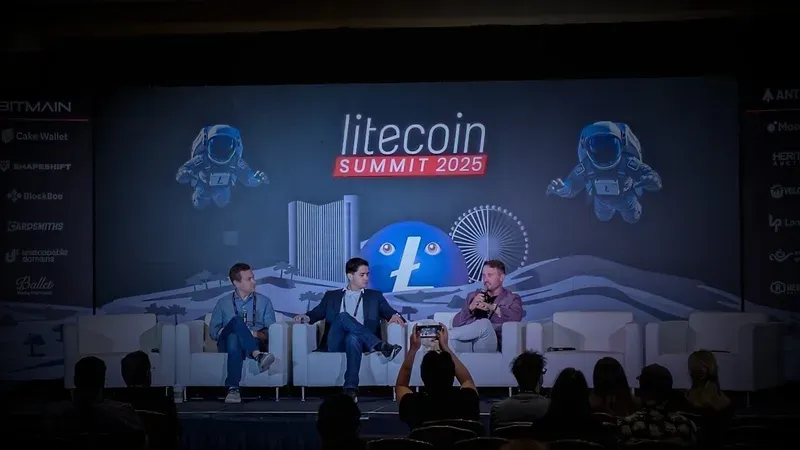 Three men seated in white chairs on stage during Litecoin Summit 2025, with a large screen behind them displaying the event title, floating astronaut mascots, and sponsor logos. One speaker holds a microphone and gestures as the audience records the panel.